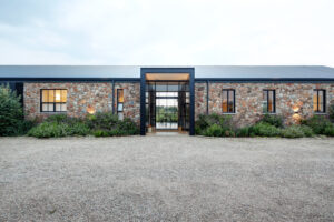 Symmetric front view of a modern stone house featuring a minimalist black metal entrance frame, large glass windows, and a wide gravel driveway.
