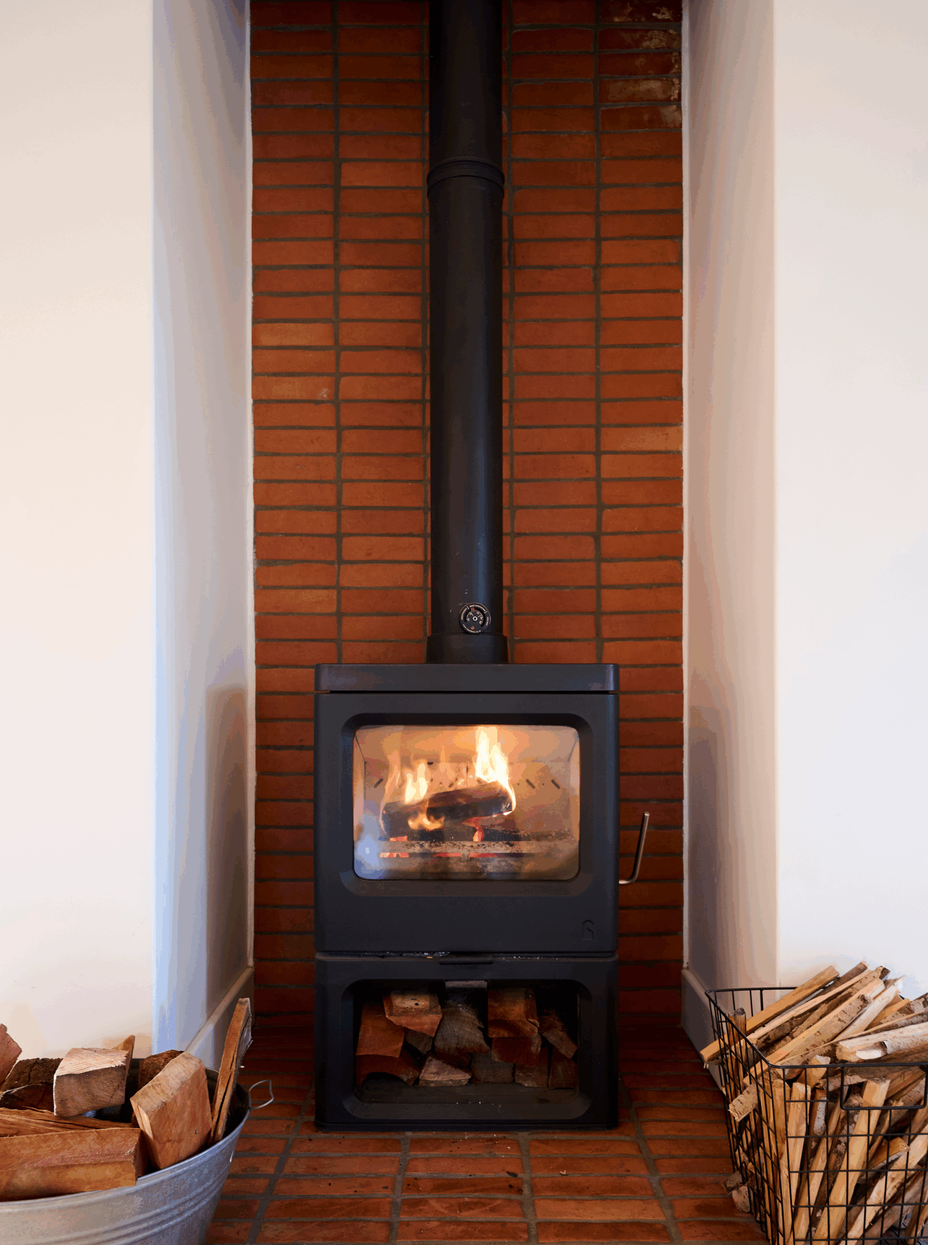 A black cast-iron wood-burning stove set against a red brick accent wall, flanked by metal bins filled with firewood.