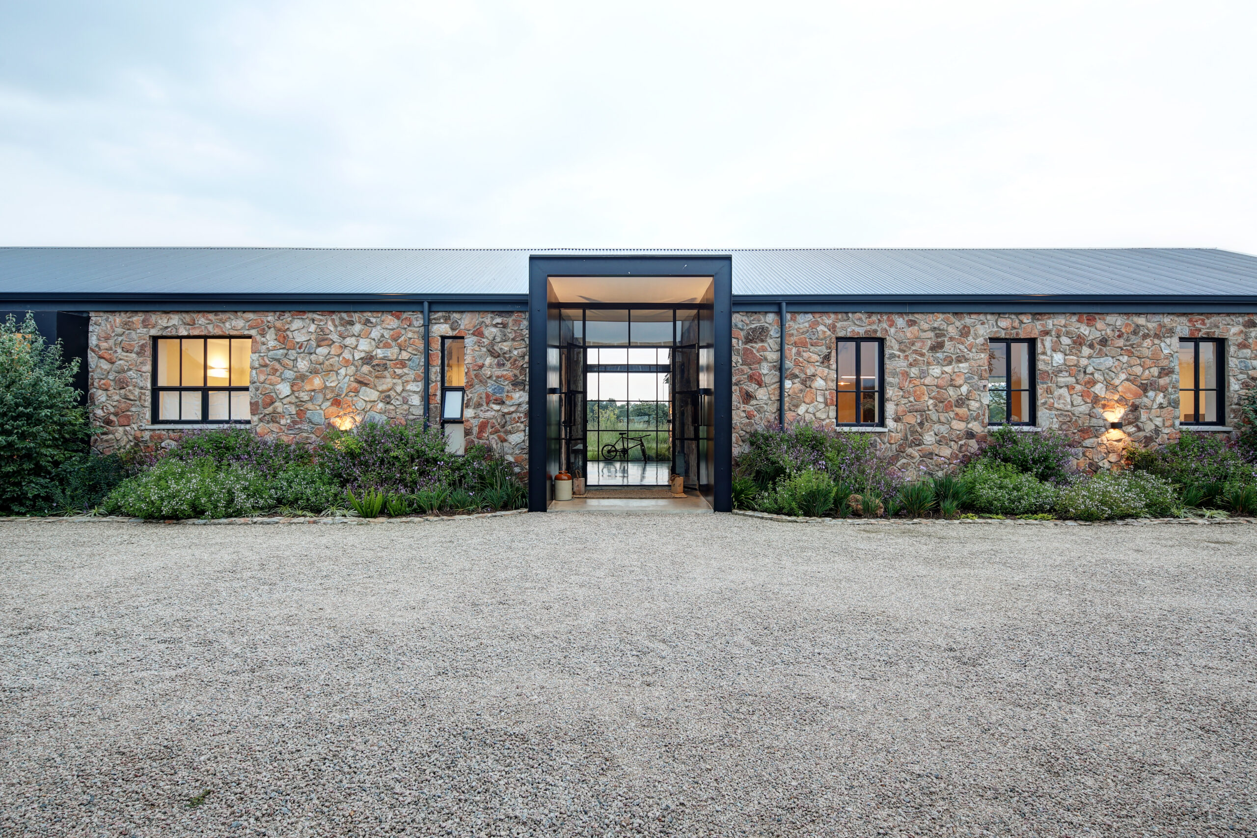 side-view-of-front-entrance Symmetric front view of a modern stone house featuring a minimalist black metal entrance frame, large glass windows, and a wide gravel driveway.