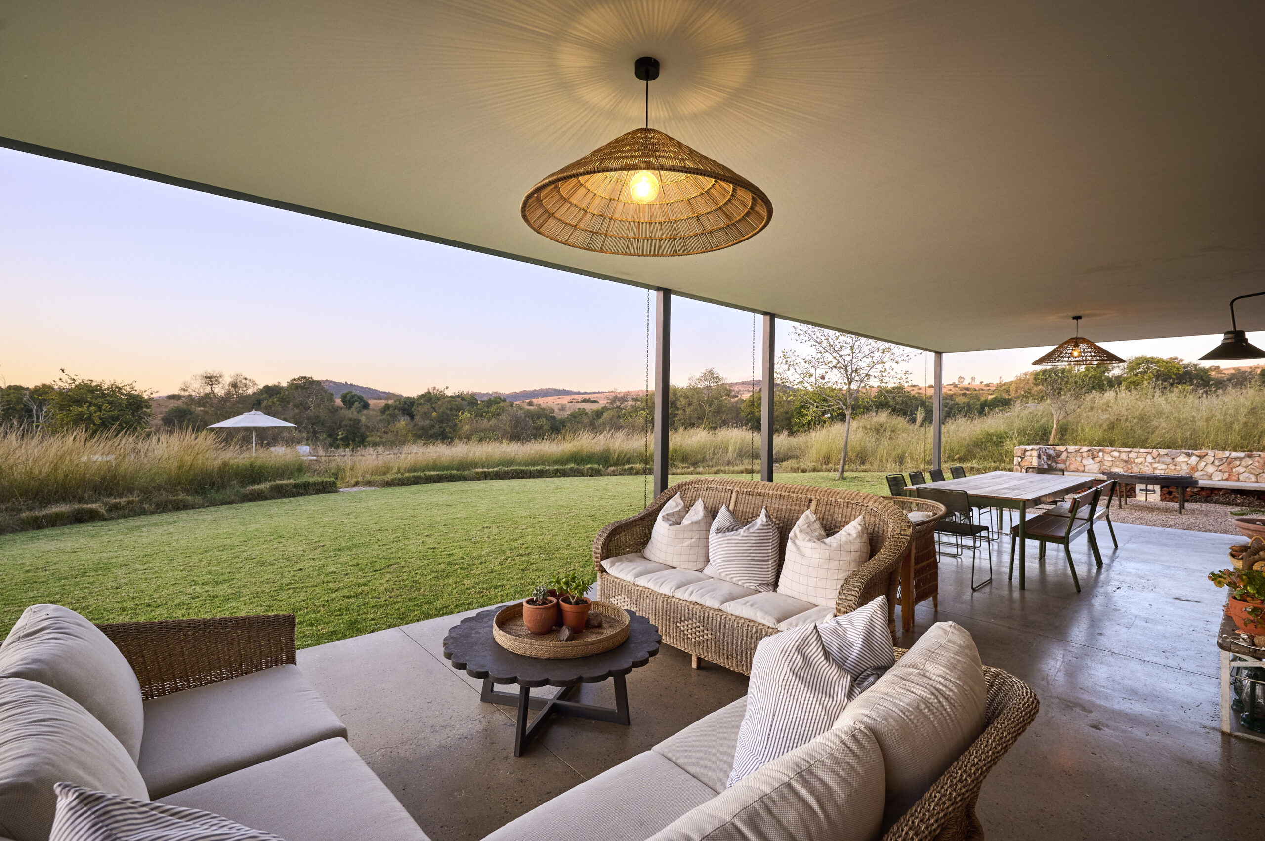 Large covered patio configured as an outdoor living room with wicker sofas, a woven pendant light, and a view of a manicured green lawn.