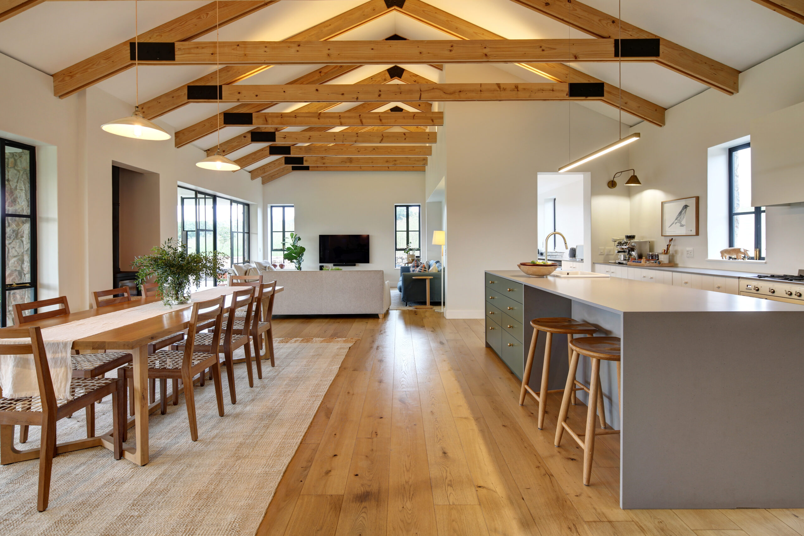 High-angle view of a large open-concept great room combining the kitchen, dining table, and living space under a vaulted timber roof.