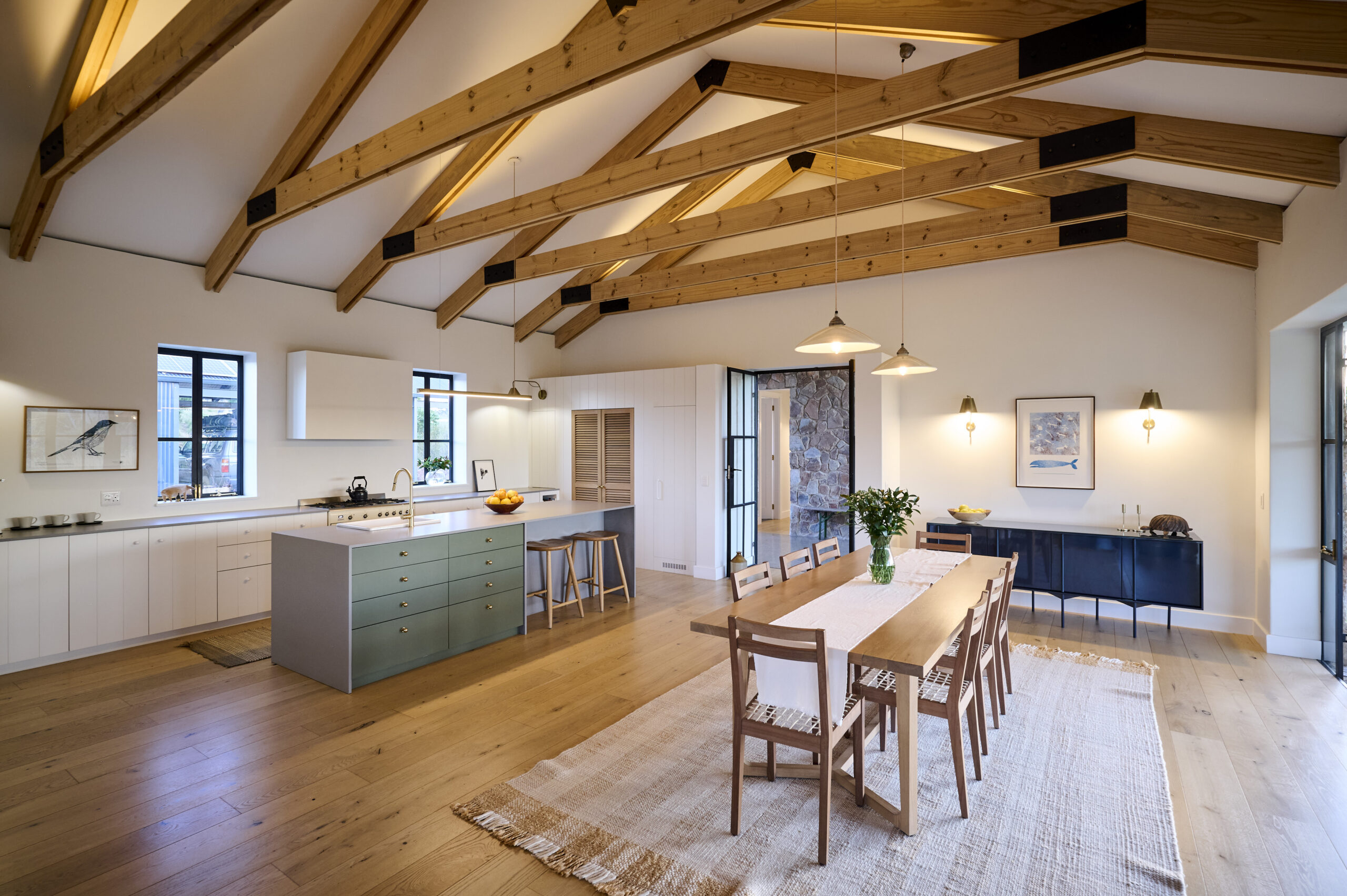 side-view-of-front-entrance Large open-plan great room with exposed wooden ceiling trusses, a modern sage green kitchen island, a long wooden dining table, and chic brass wall sconces.