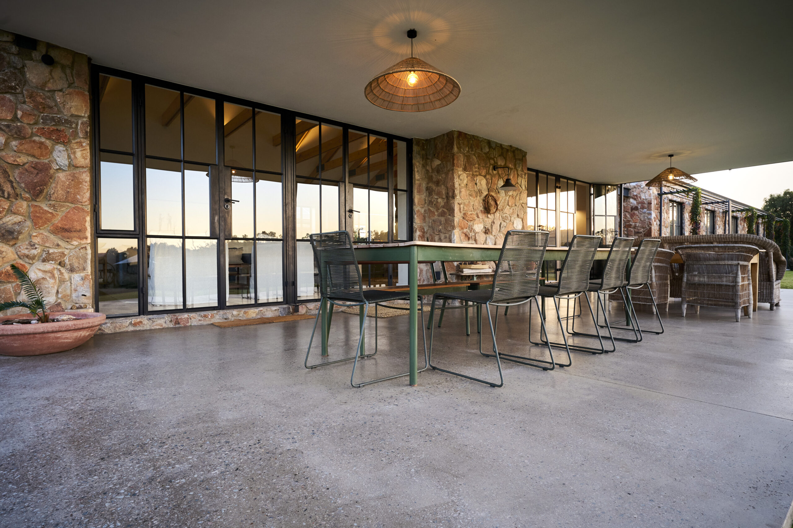 Long outdoor dining table with metal chairs on a polished concrete patio floor beneath a covered roof extension.