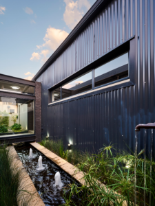 Close up of a linear pond with three fountains set in a narrow courtyard between black facebrick and corrugated iron walls.