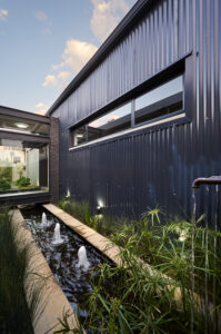 Close-up of the linear pond and fountains at dusk, with warm lighting reflecting off the black corrugated iron.