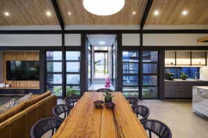 Open-plan dining room with a long solid wood table, framed by black portal steel beams and views of the internal courtyard.