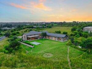 High-angle drone aerial view of the modern grey house, showcasing the sprawling green lawns, swimming pool, and surrounding countryside.