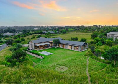 High-angle drone aerial view of the modern grey house, showcasing the sprawling green lawns, swimming pool, and surrounding countryside.