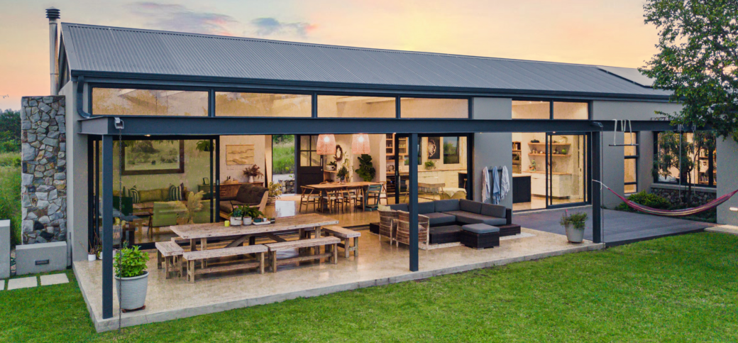 Full exterior view of a luxury modern farmhouse at dusk, showing a large covered patio with an outdoor dining table, lounge set, and seamless glass sliding doors opening to the interior.