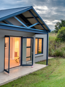 Exterior view of a modern grey residential wing with open black-framed glass double doors leading out onto a green lawn.