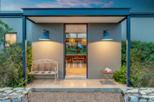 Modern home front entrance at dusk with symmetric blue wall sconces illuminating a grey exterior and a view into the warm dining room.