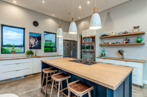 Modern kitchen featuring a large wooden-top island with a built-in stove top, wooden bar stools, white cabinetry, open floating shelves, and chic white pendant lights.
