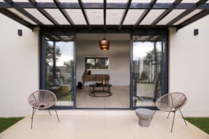A symmetrical shot of a covered outdoor terrace with two modern chairs facing a glass door under a pergola-style roof.