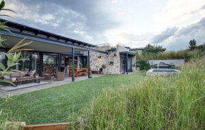 A wide-angle view of a modern farmhouse exterior featuring an open-plan outdoor living area, stone masonry walls, a green lawn, and a small rectangular swimming pool under a cloudy sky.