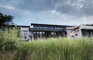 Low-angle view of the back of a modern house with large glass windows and an outdoor living area overlooking a green field.