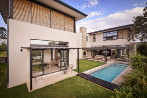 Wide shot of a luxury backyard featuring an L-shaped swimming pool, green lawn, and a covered patio area.