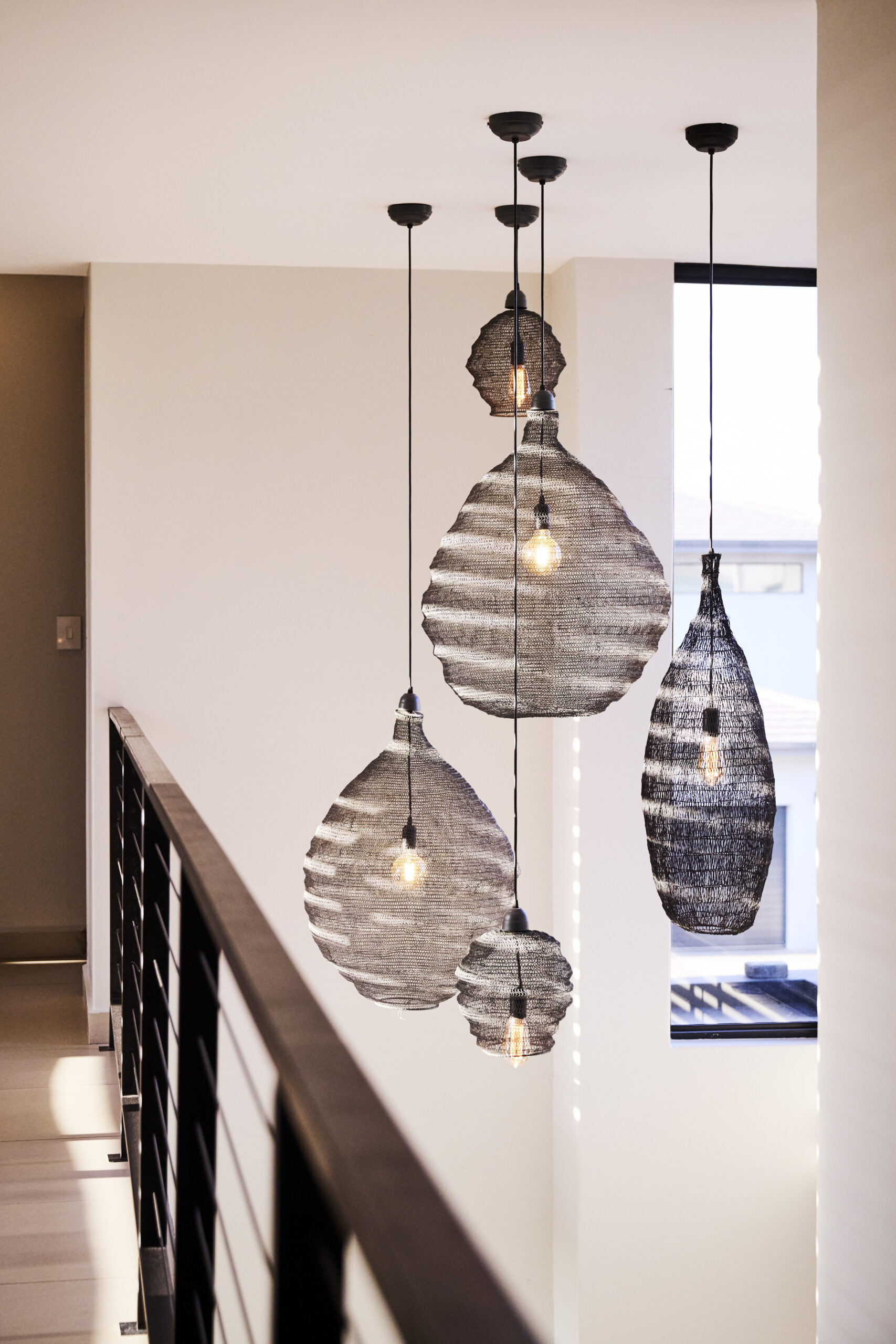 modern-staircase-pendant-lighting A split image showing a glass-enclosed staircase and a cluster of large, black woven pendant lights hanging in a void.