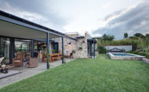 A modern patio design with a wooden dining table, wicker seating, and an integrated outdoor kitchen, adjacent to a stone-walled pool area and manicured lawn.