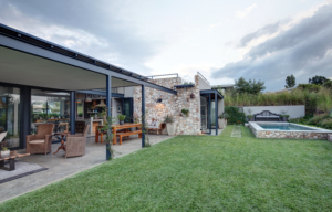 A modern patio design with a wooden dining table, wicker seating, and an integrated outdoor kitchen, adjacent to a stone-walled pool area and manicured lawn.