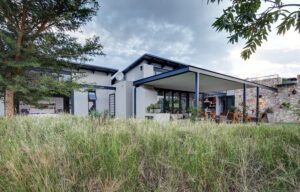 Outdoor patio with a wooden dining table, built-in kitchen, and a covered lounge area overlooking a lawn.