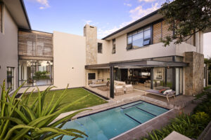 Wide shot of a luxury backyard featuring an L-shaped swimming pool, green lawn, and a covered patio area.