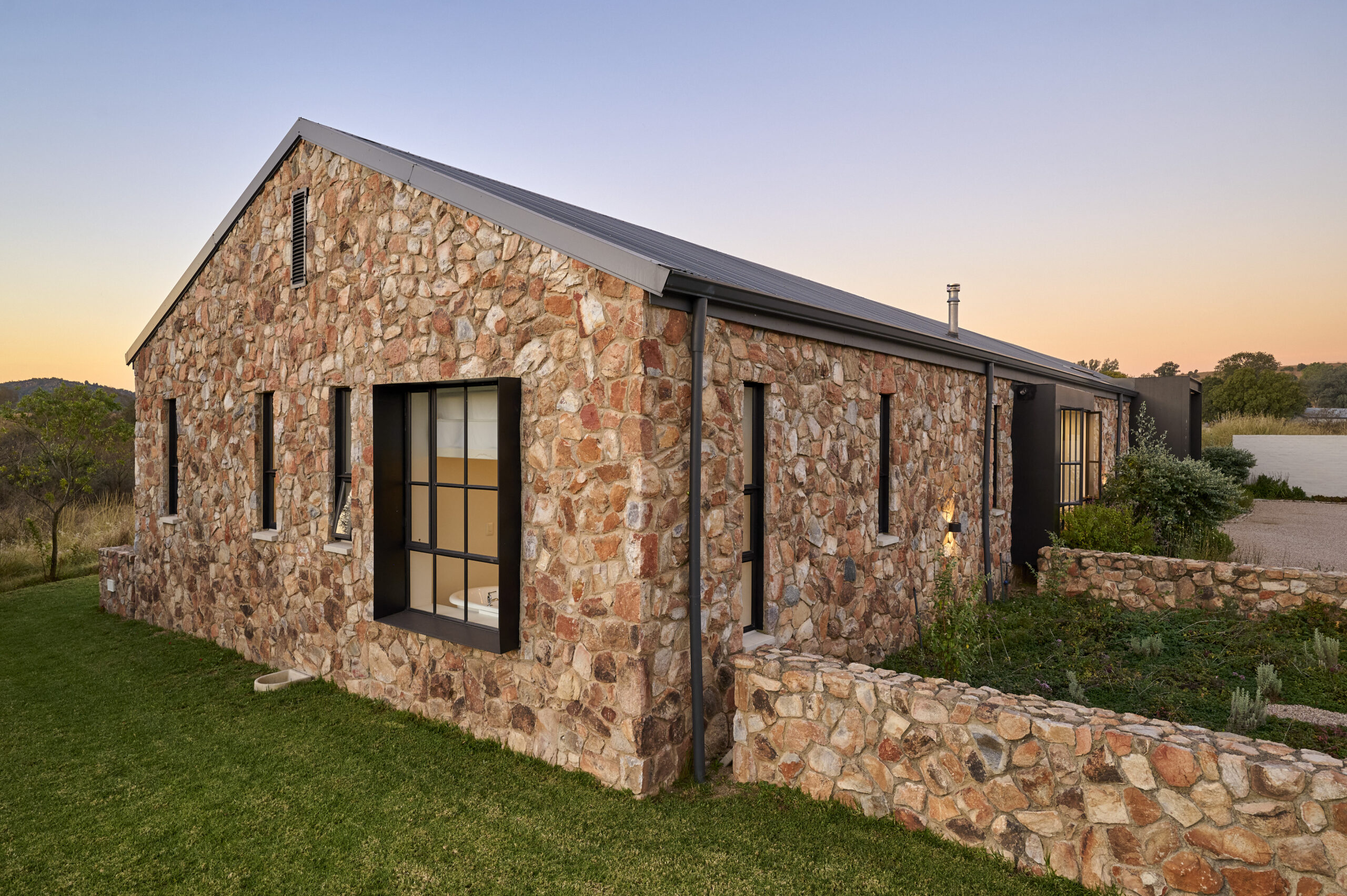 modern-stone-architecture-gable-end-roof Perspective view of the stone house's gable wall showing clean rooflines, black-framed windows, and manicured lawn.
