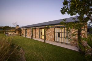 Long shot of the rear stone facade and glass doors bordering a green grass lawn at sunset.