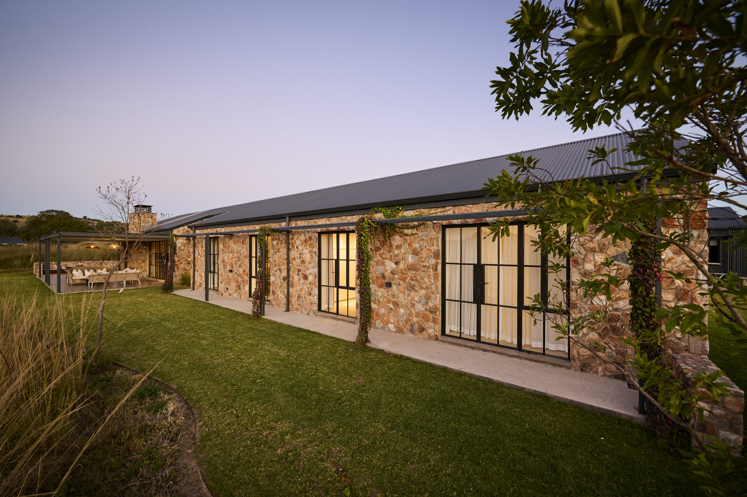 Long shot of the rear stone facade and glass doors bordering a green grass lawn at sunset.