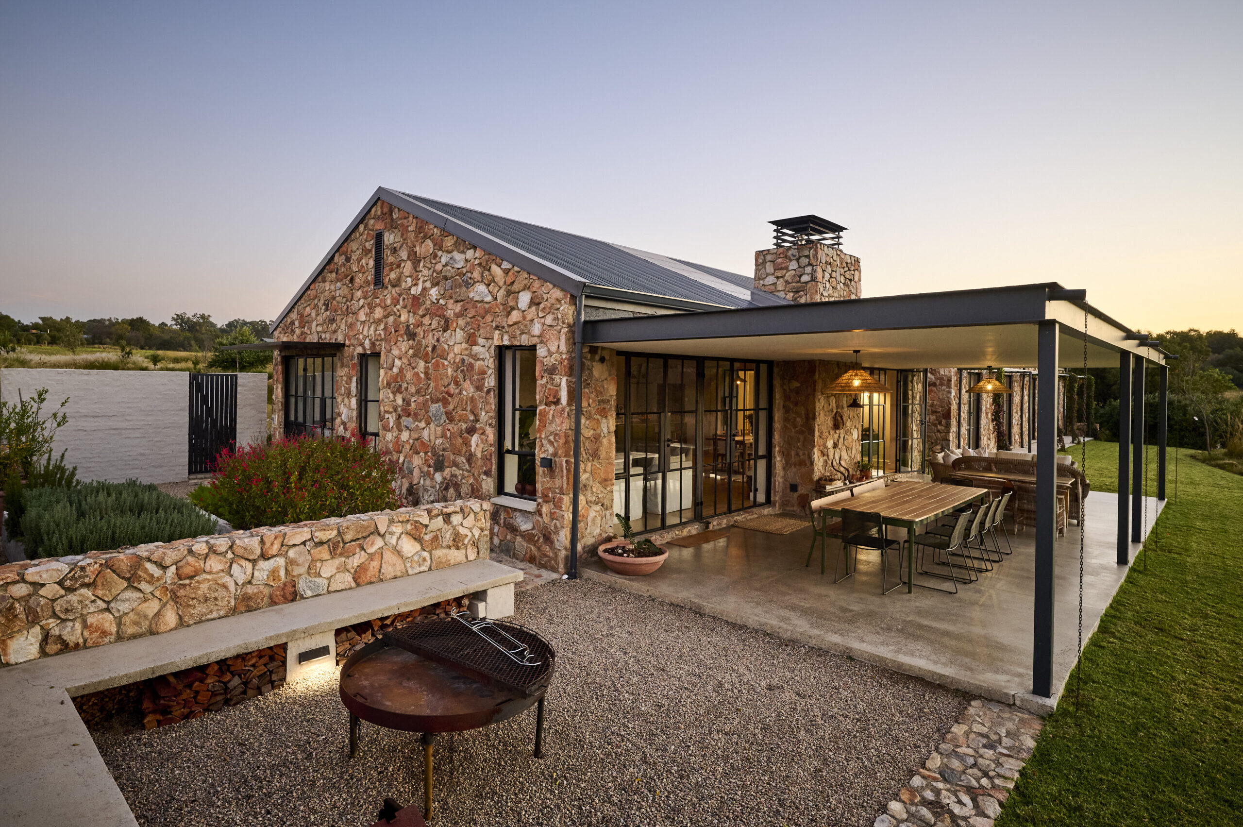 side-view-of-front-entrance Rear exterior of the stone house showing a covered veranda, an outdoor dining setup, and a gravel boma area with a built-in stone bench and fire pit.