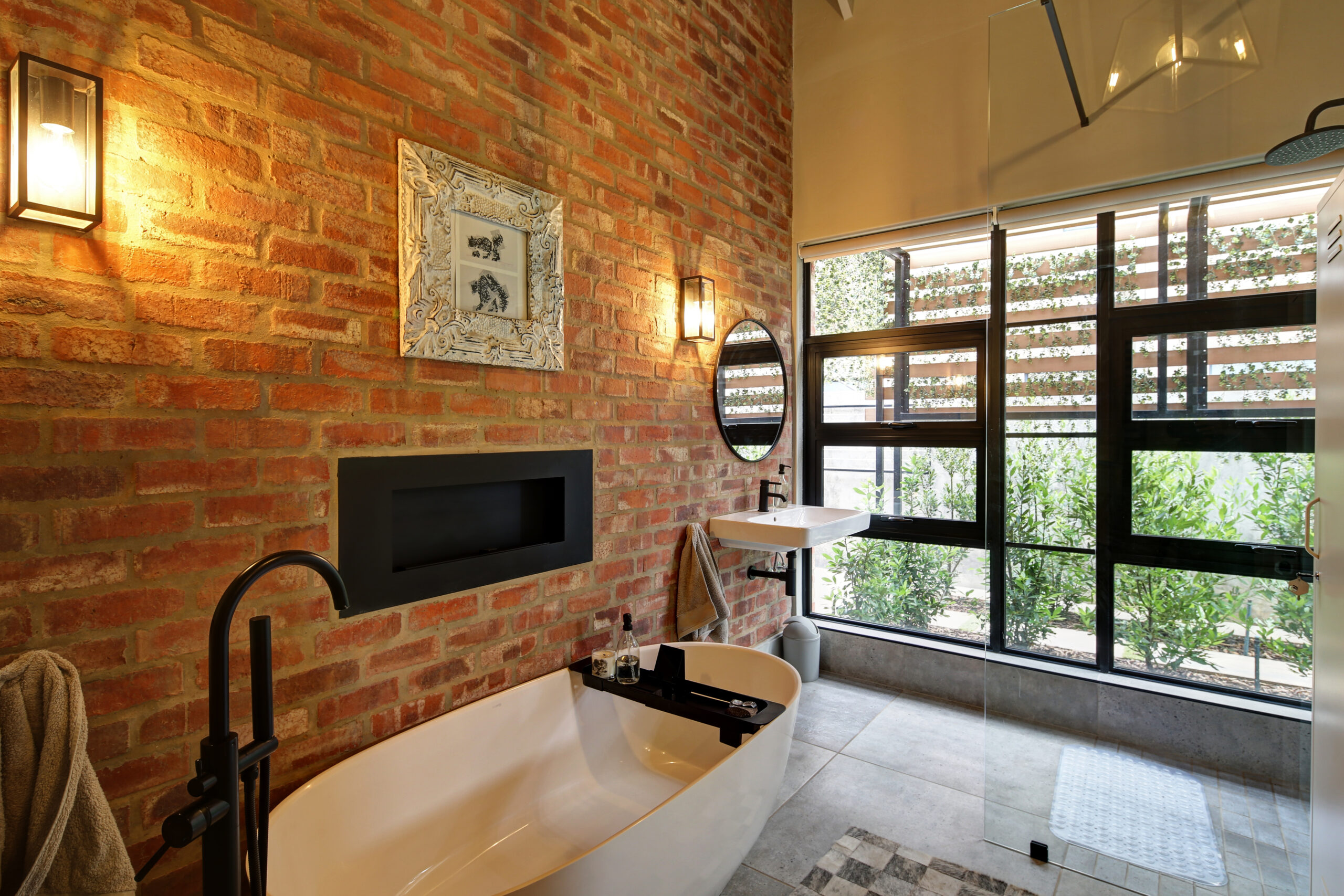 En-suite bathroom with exposed stock brick wall looking out toward timber and steel pergola and courtyard garden